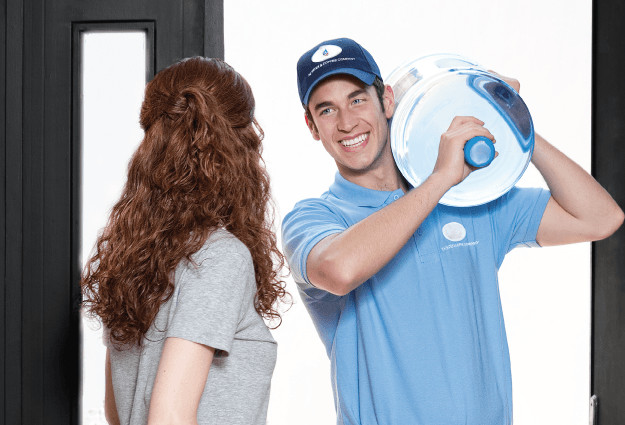 На фото радісний доставник води Aqua Plus приносить воду в квартиру, його зустрічає дівчина, яка замовила воду
